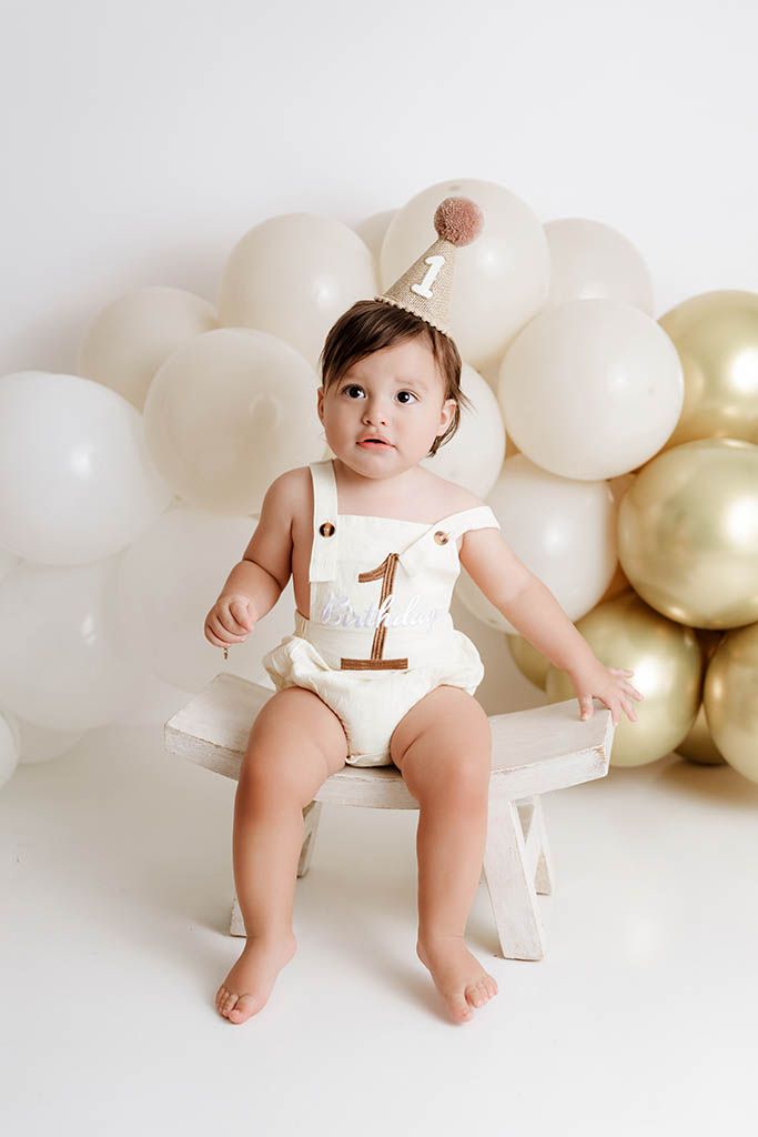 One year old baby sitting on white bench wearing cream romper and party hat by Temescal Valley baby photographer.