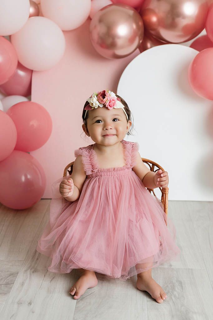 Smiling baby girl in pink tulle dress and floral headband standing among pink and rose gold balloons.