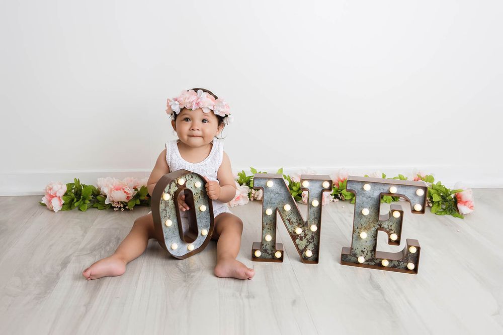 Baby girl in floral crown sits beside marquee ONE letters with pink flowers for first birthday.