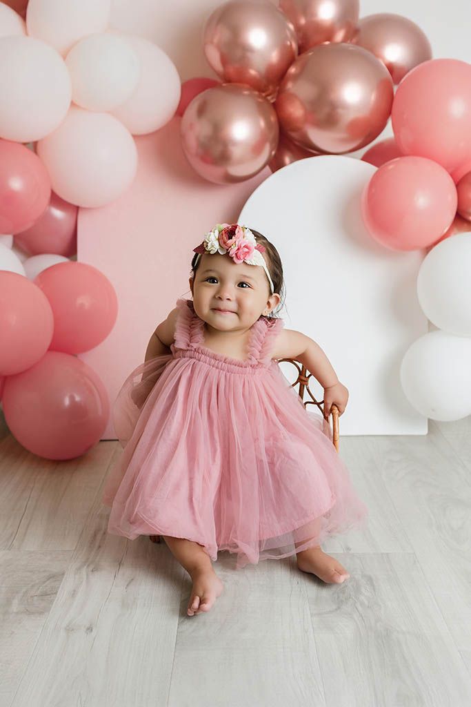 A baby girl in a pink tulle dress and floral headband sits surrounded by pink and rose gold balloons by Perris baby photographer.