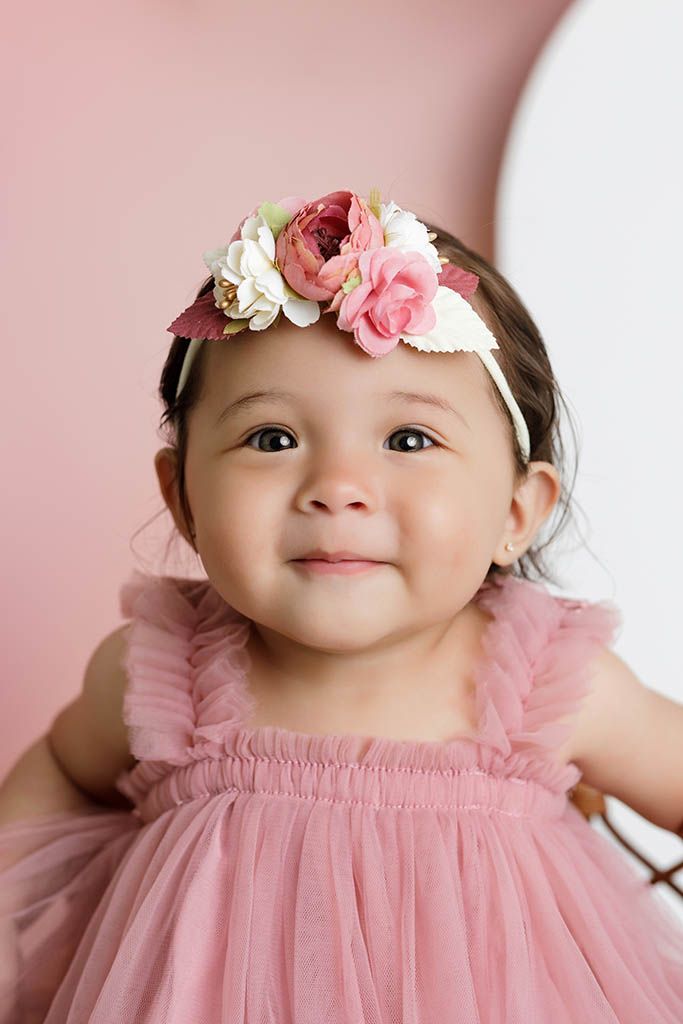 Adorable baby girl in pink tulle dress and floral headband smiling sweetly against a soft pink background.