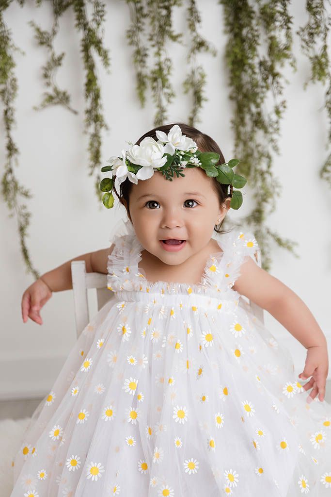 Smiling baby girl in white daisy dress and floral crown poses for portrait photo with greenery backdrop.