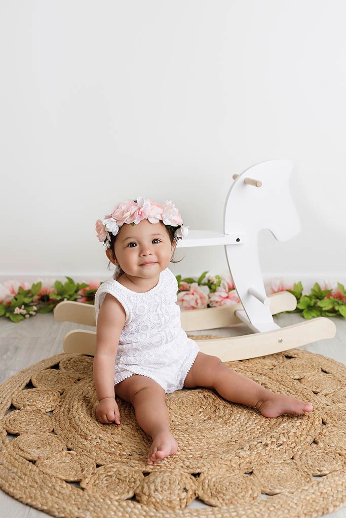 Smiling baby girl in white romper and floral crown sits on jute rug beside white wooden rocking horse.