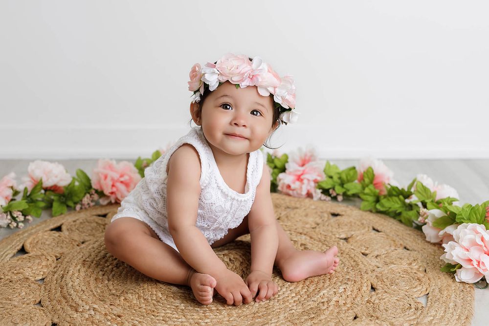 Baby girl in white romper and floral crown sits on woven rug surrounded by pink flowers.