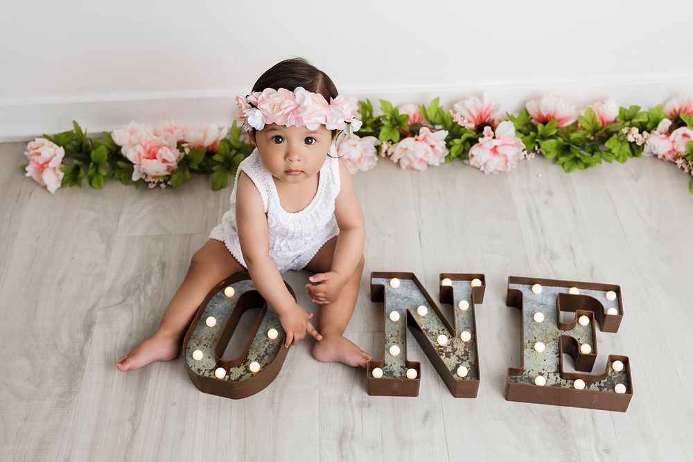 Baby girl in white dress and floral crown sits beside marquee ONE letters by Perris baby photographer