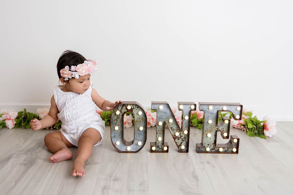 Baby girl in white romper sits beside lit marquee ONE letters with pink flowers for first birthday photo by Perris baby photographer.