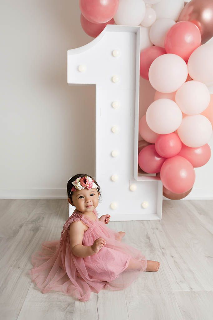 A baby girl in a pink tutu dress sits in front of a large lit number 1 with pink balloons for her first birthday by Perris baby photographer