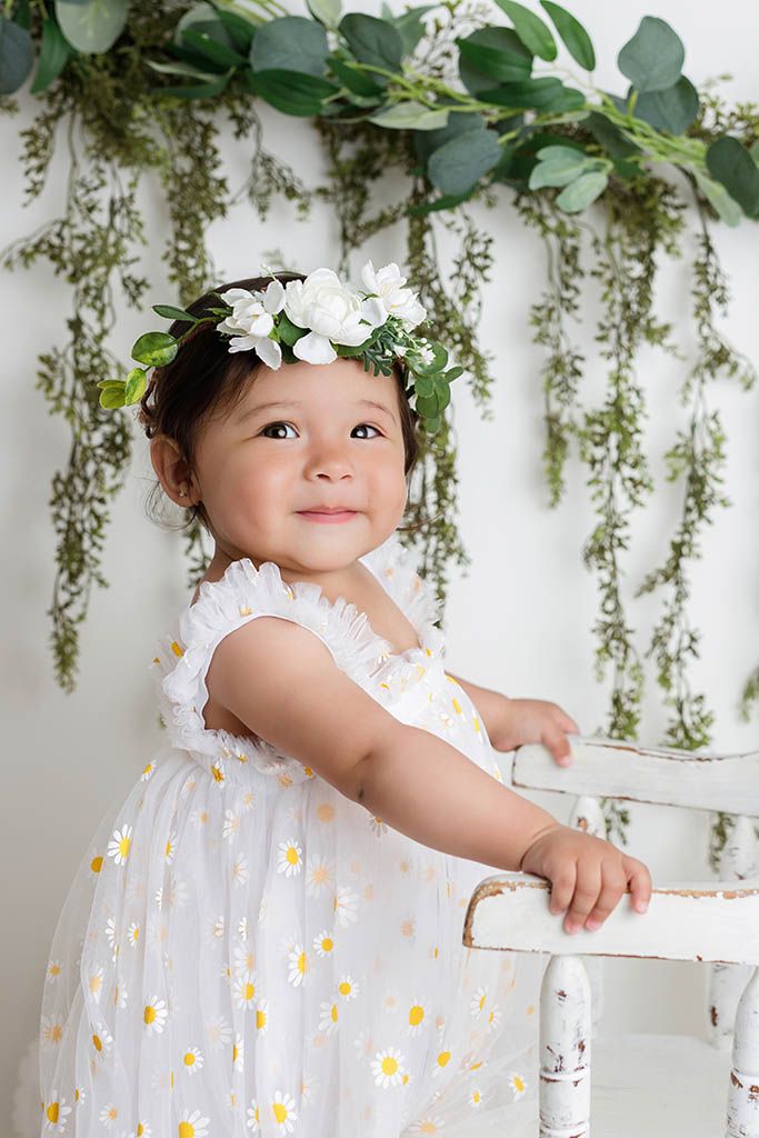 Toddler girl in daisy dress and floral crown smiles sweetly against a white backdrop with green garland.