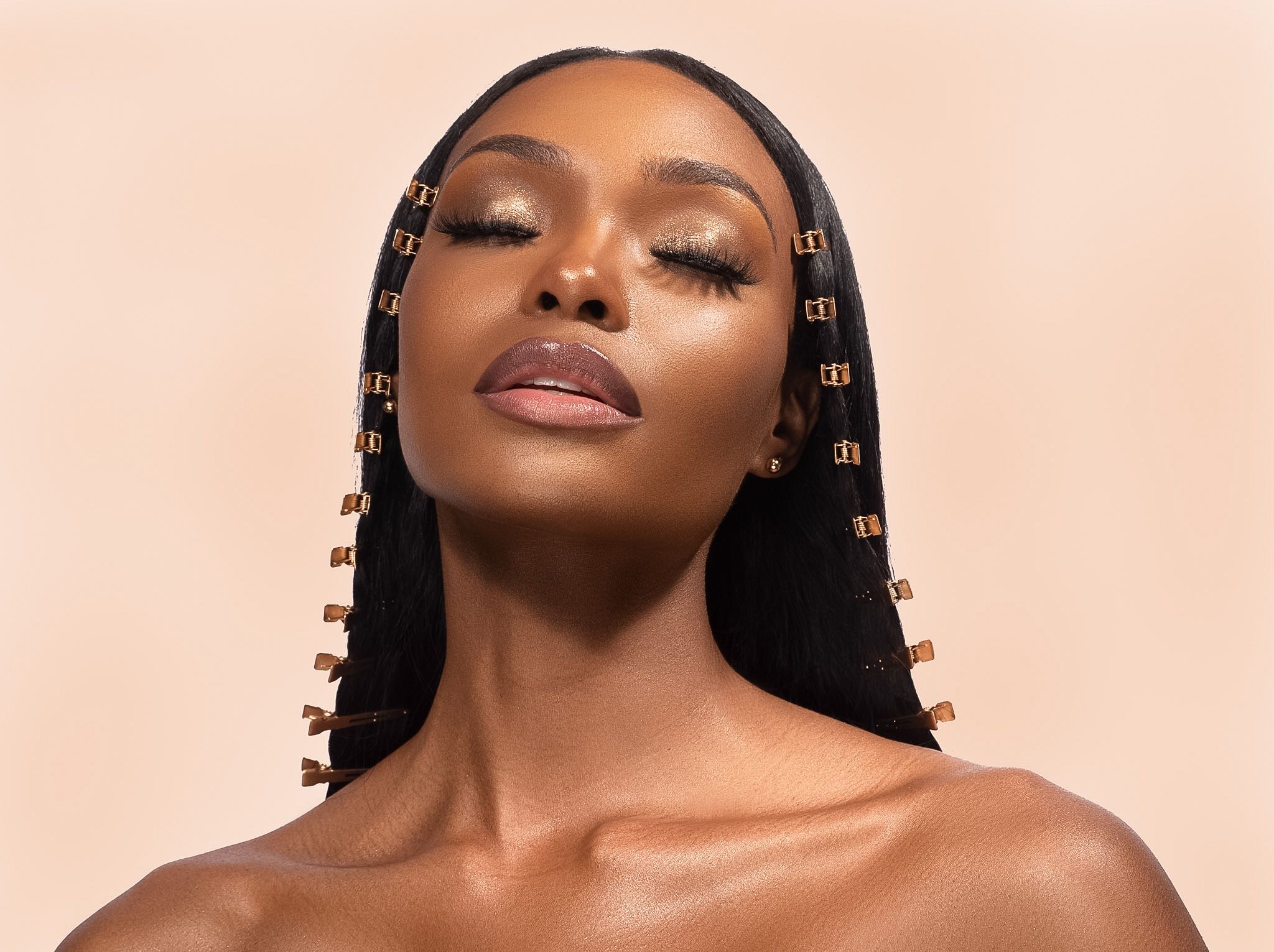 A woman with long braids adorned with gold clips poses against a beige background, wearing elegant makeup.