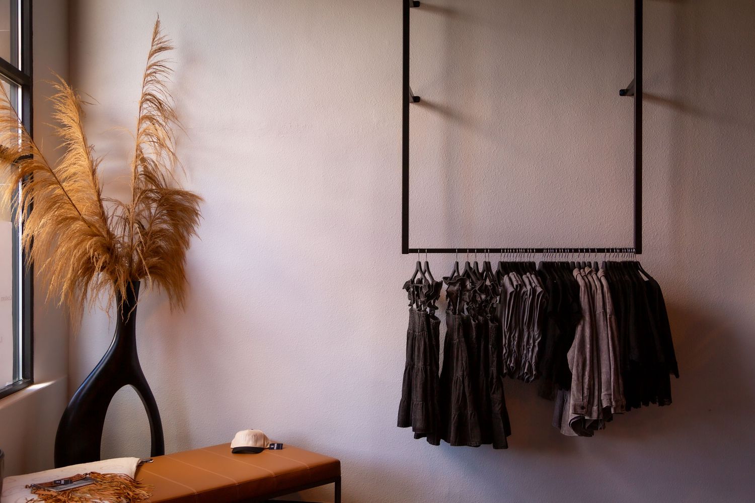 Minimalist wardrobe rack with dark clothing hangs on wall beside pampas grass in black vase and leather suitcase.