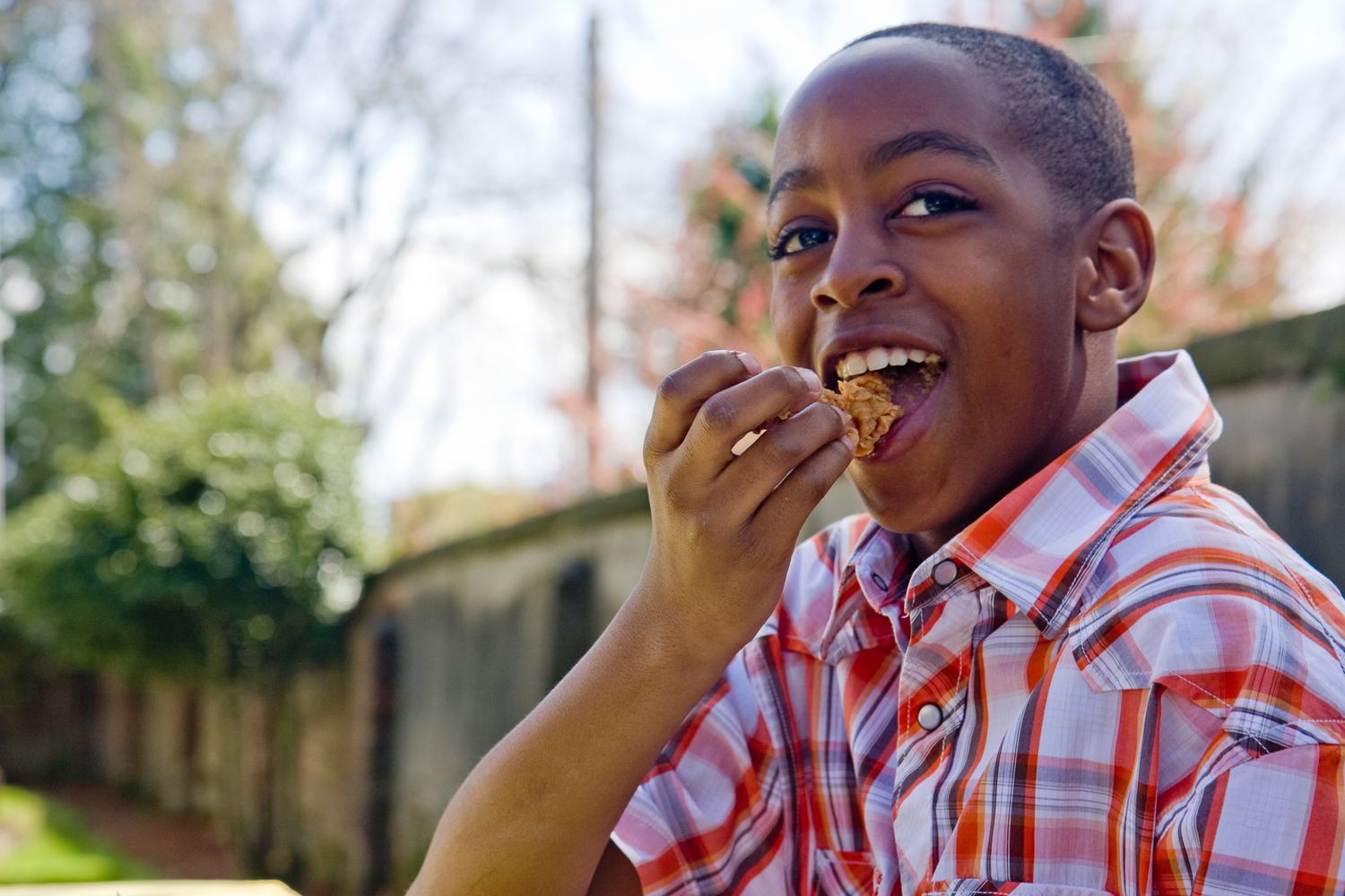 Child wearing an orange plaid shirt takes a joyful bite of food outdoors in a backyard setting.