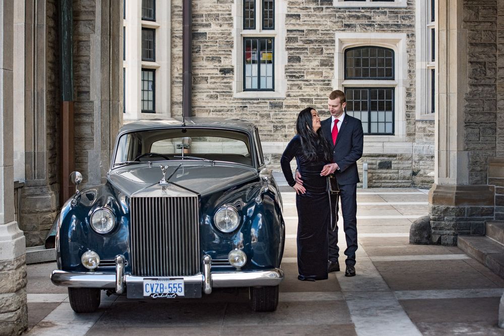 Elegant couple standing beside a vintage Rolls-Royce outside a grand stone building.