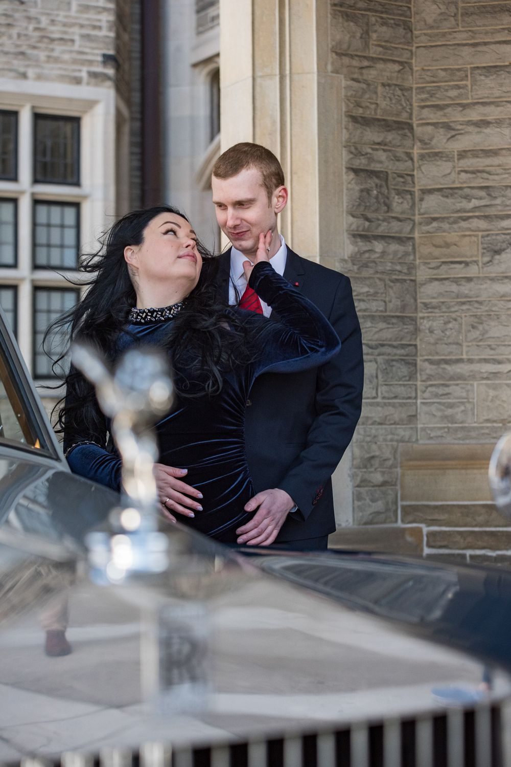 Couple embracing near a classic Rolls-Royce in front of a historic stone building.