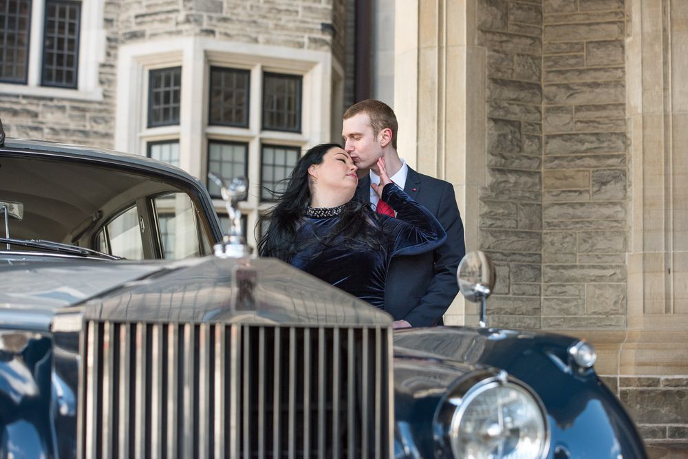 Couple sharing a kiss near a classic Rolls-Royce in front of a stone building.