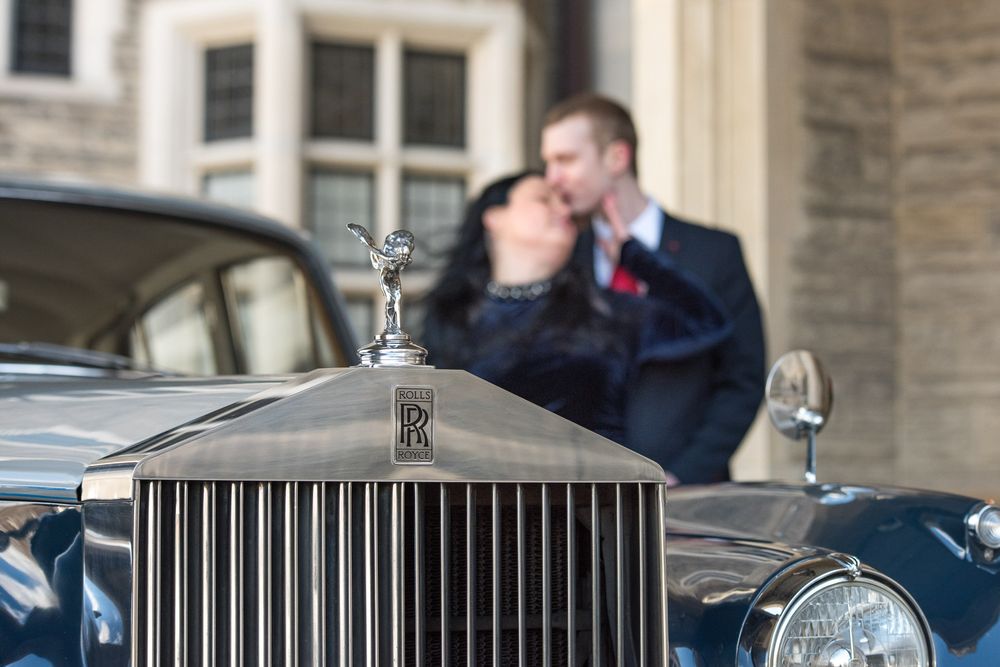 Couple kissing in the background with a vintage Rolls-Royce hood ornament in focus.