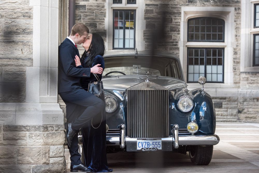 Couple sharing a kiss leaning against a classic vintage Rolls Royce outside a stone building.