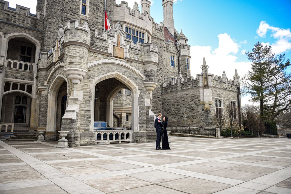 Couple posing in front of a majestic Gothic stone castle with Canadian flags flying.