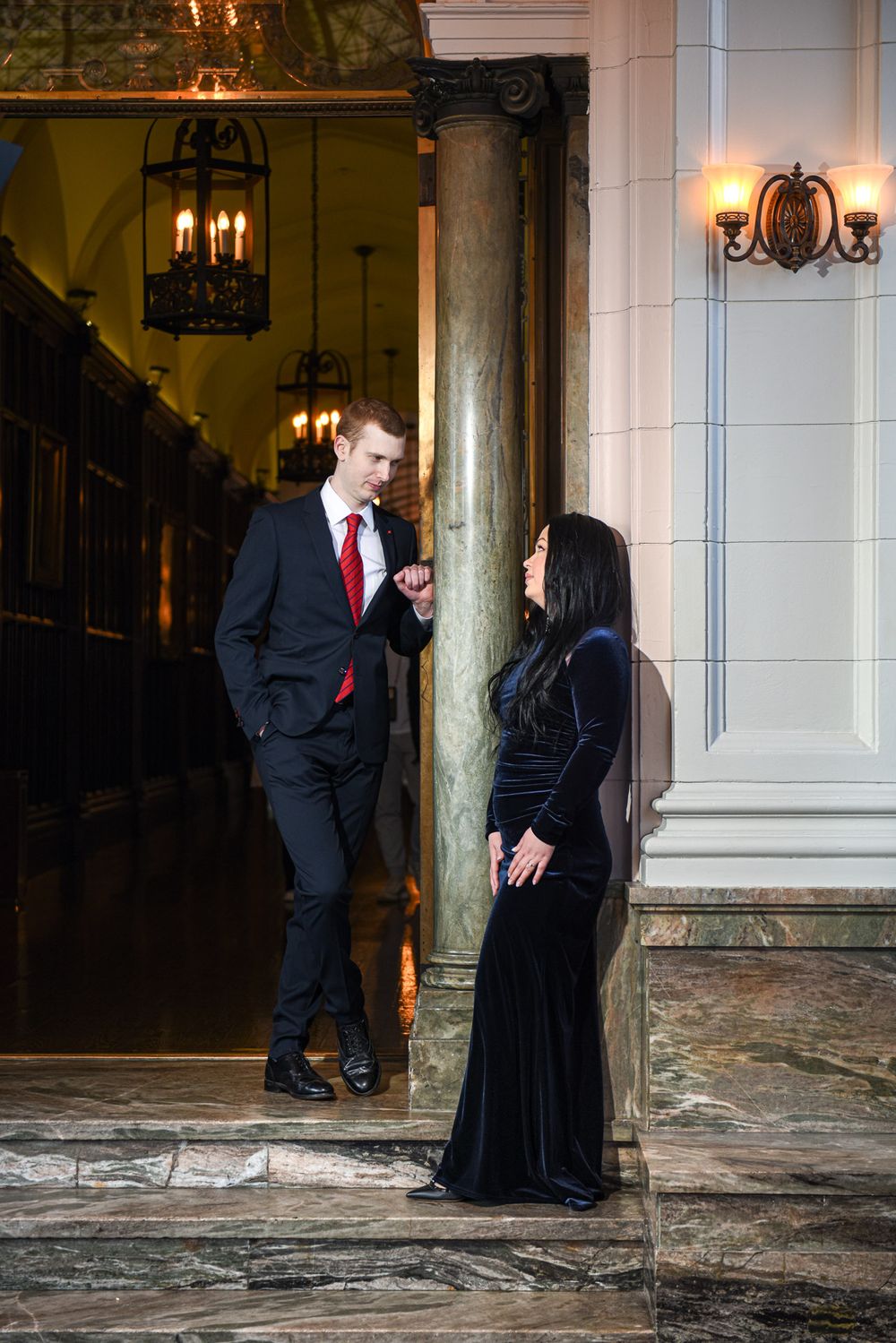 Elegant couple in black formal wear conversing by marble column in a grand ornate hall.