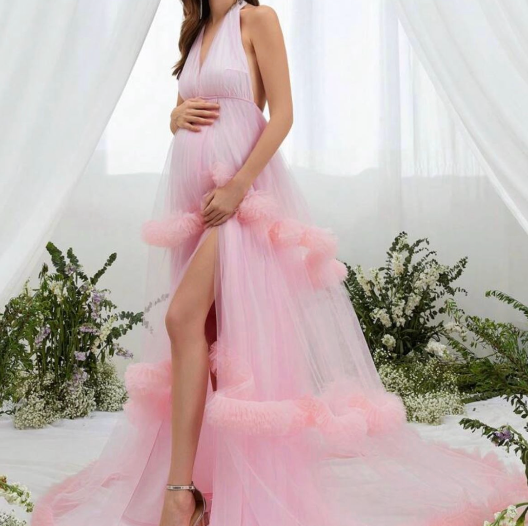 Pregnant woman in a pink tulle halter gown with ruffled hem and thigh slit, posing in a floral garden setting.