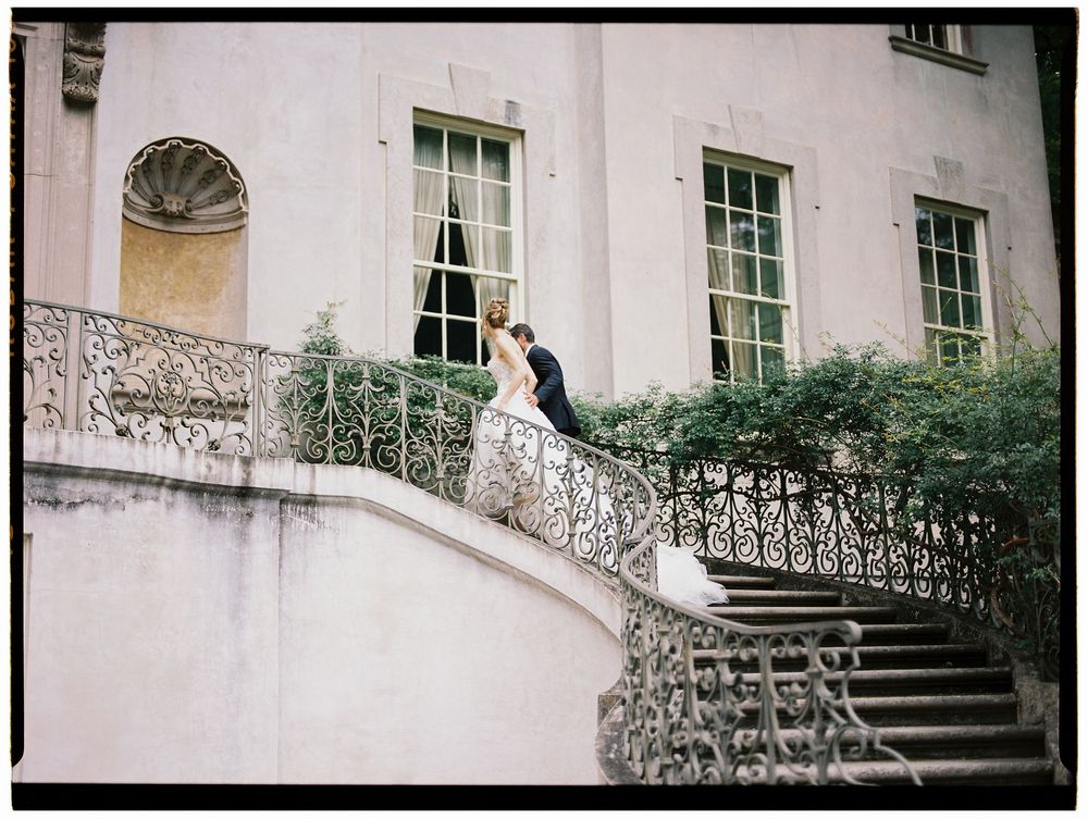 Bride and groom share a romantic moment on ornate iron staircase of elegant white mansion during wedding photo session.