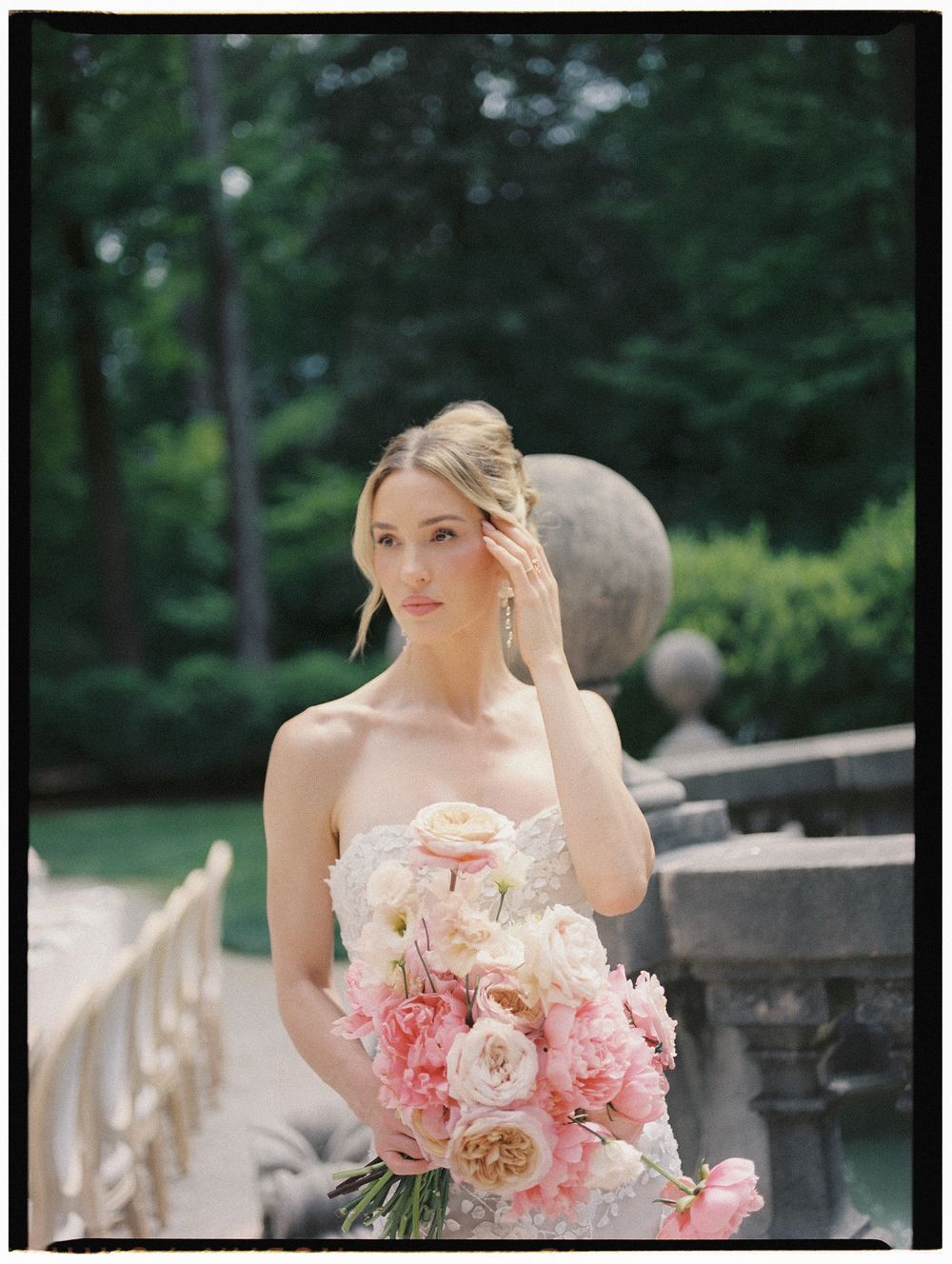 Elegant bride in strapless lace gown holding pink floral bouquet in a garden setting.