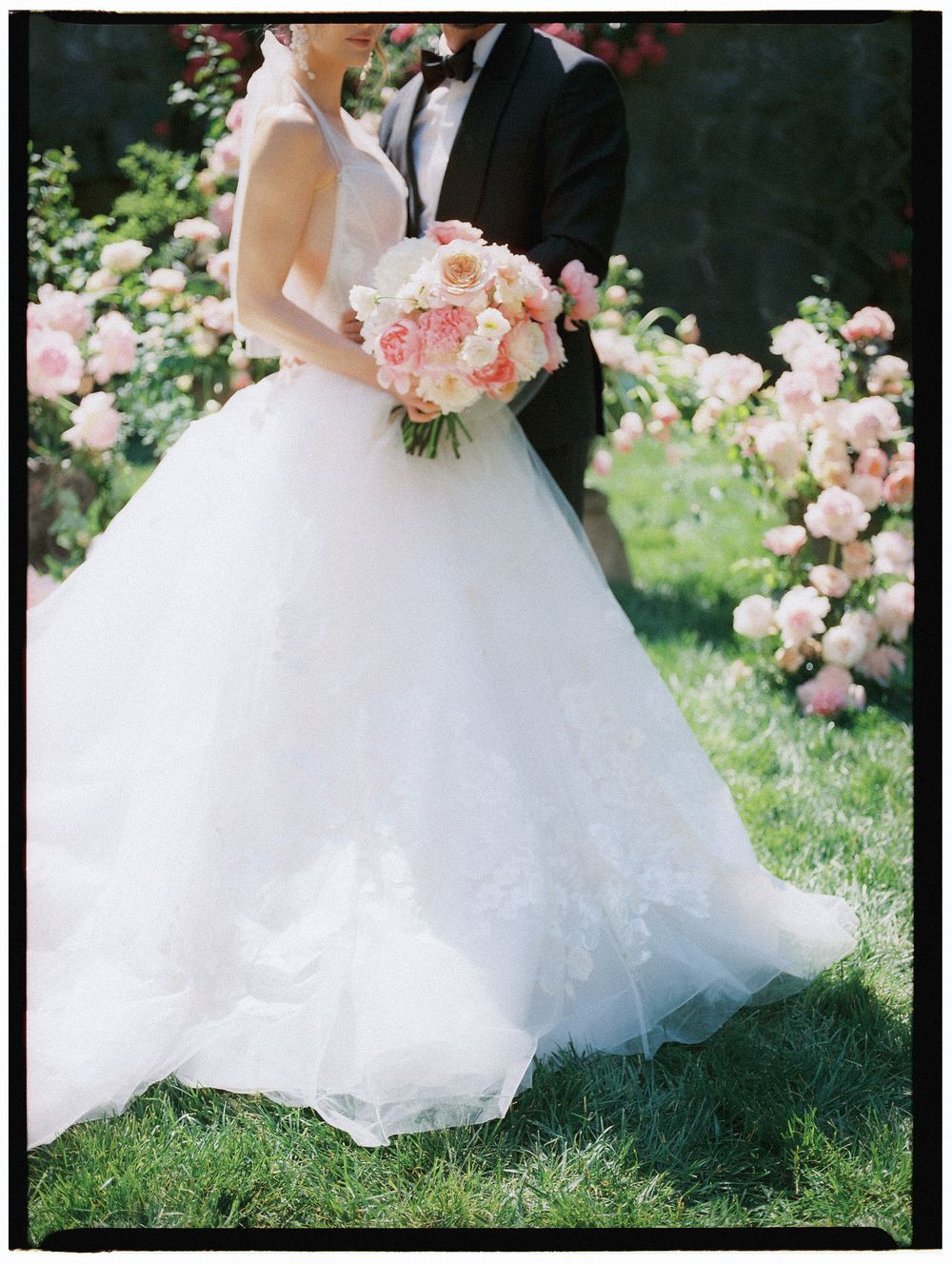 Bride in a flowing white lace ballgown holds a pink rose bouquet beside groom in black tuxedo among garden roses.