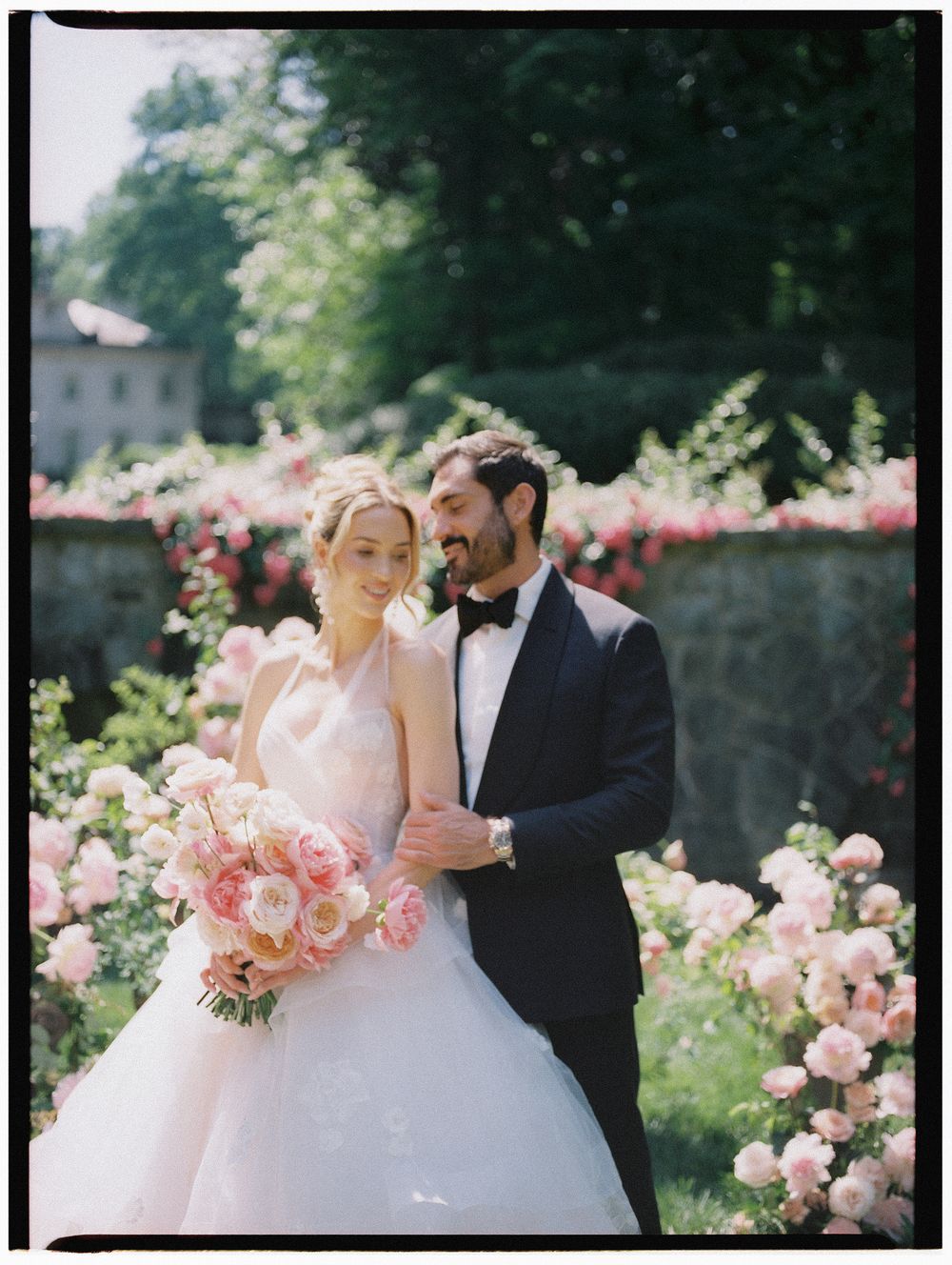 Bride and groom embrace among pink roses, she holds a blush bouquet in a white ballgown, he wears a black tuxedo.