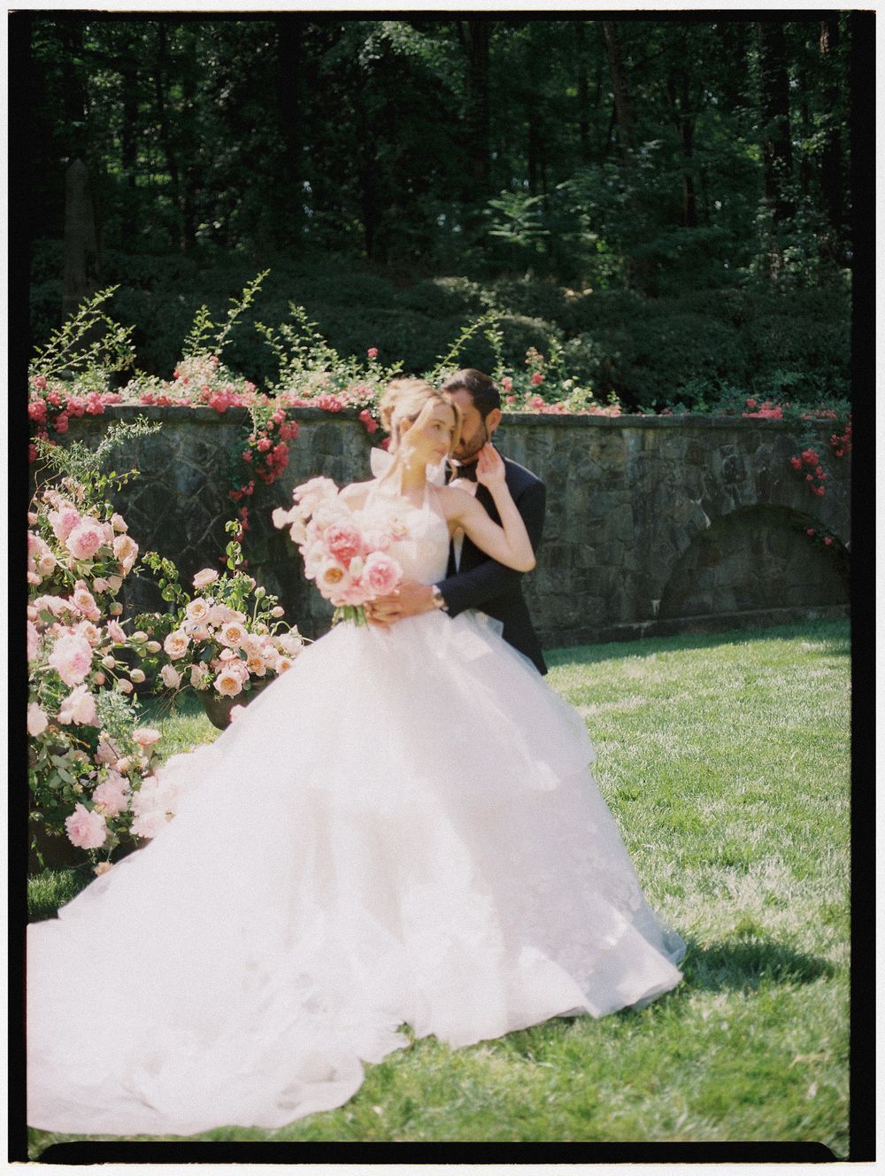 Bride and groom share a kiss in garden with pink floral arch and lush greenery backdrop.