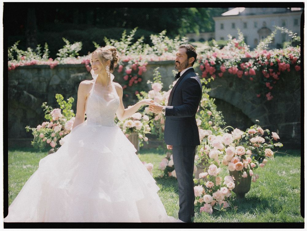 Bride in white gown and groom in navy suit exchange vows outdoors surrounded by lush pink floral arrangements.
