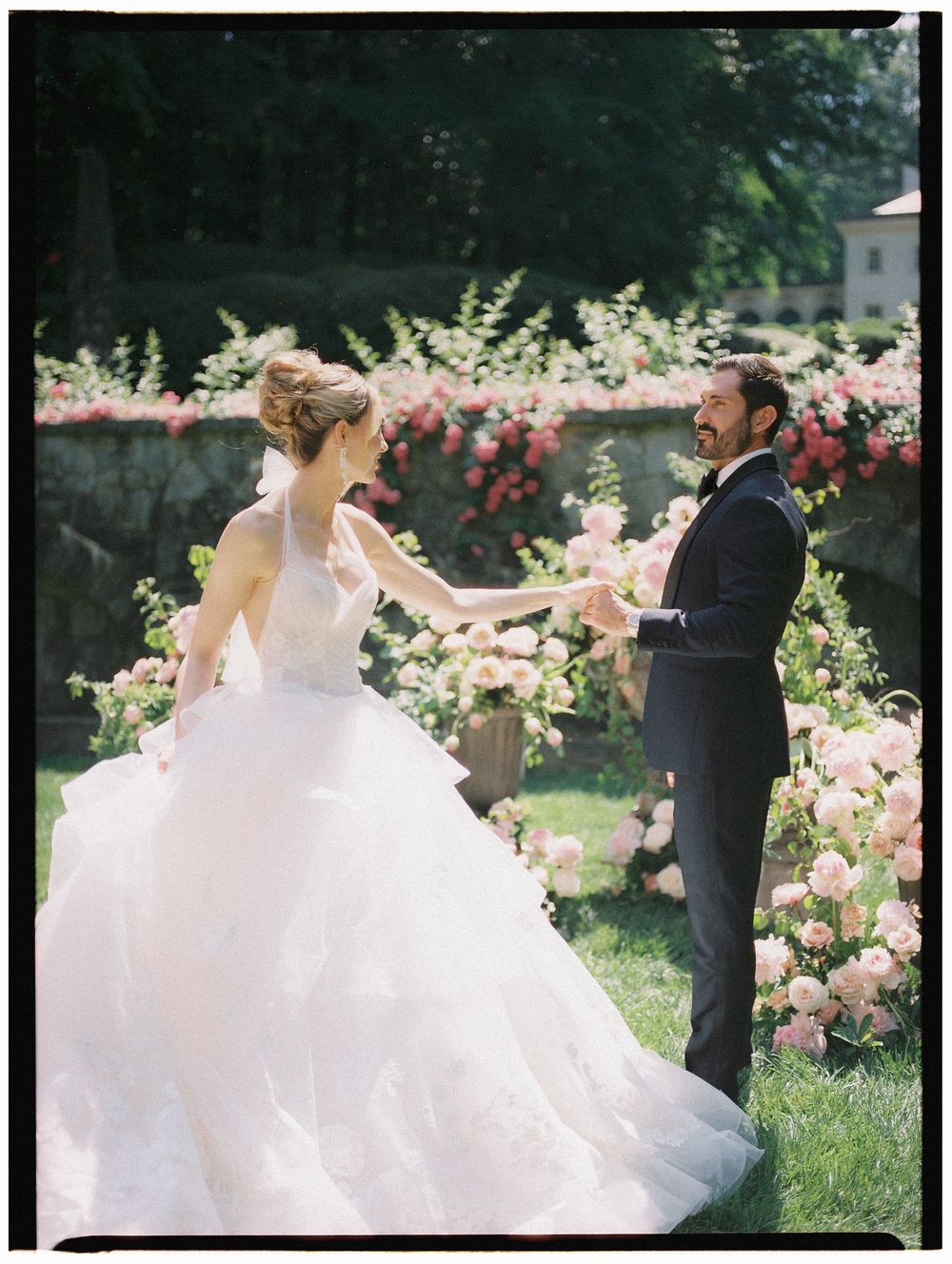 Bride and groom dancing joyfully outdoors surrounded by lush pink roses in a romantic garden wedding setting.