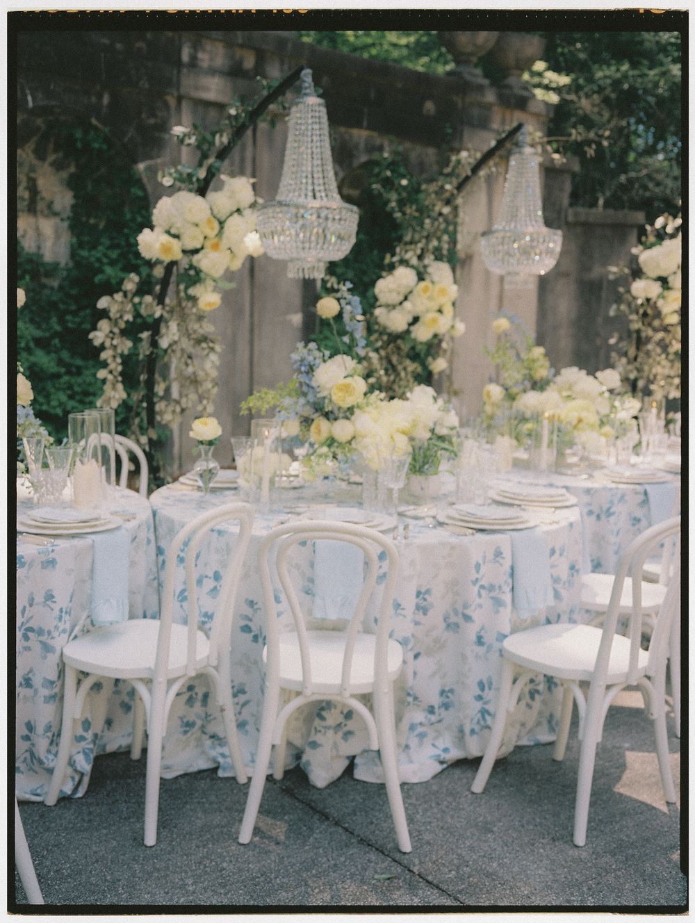 Elegant outdoor wedding reception with white floral tablecloth, crystal chandeliers, white chairs, and yellow and white floral centerpieces.