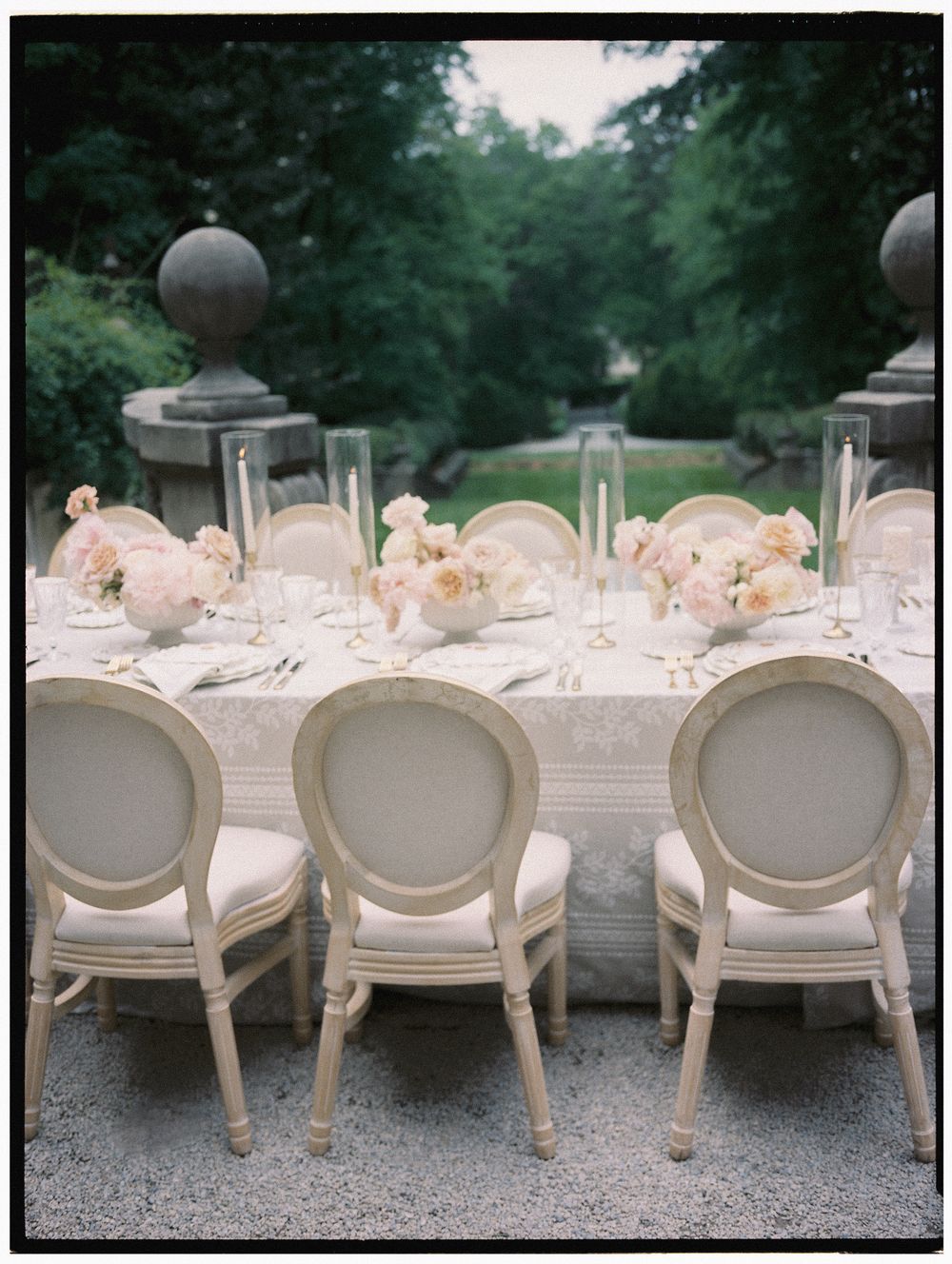 Elegant outdoor wedding reception table with ivory Louis XVI chairs, blush floral centerpieces, and crystal candle holders.