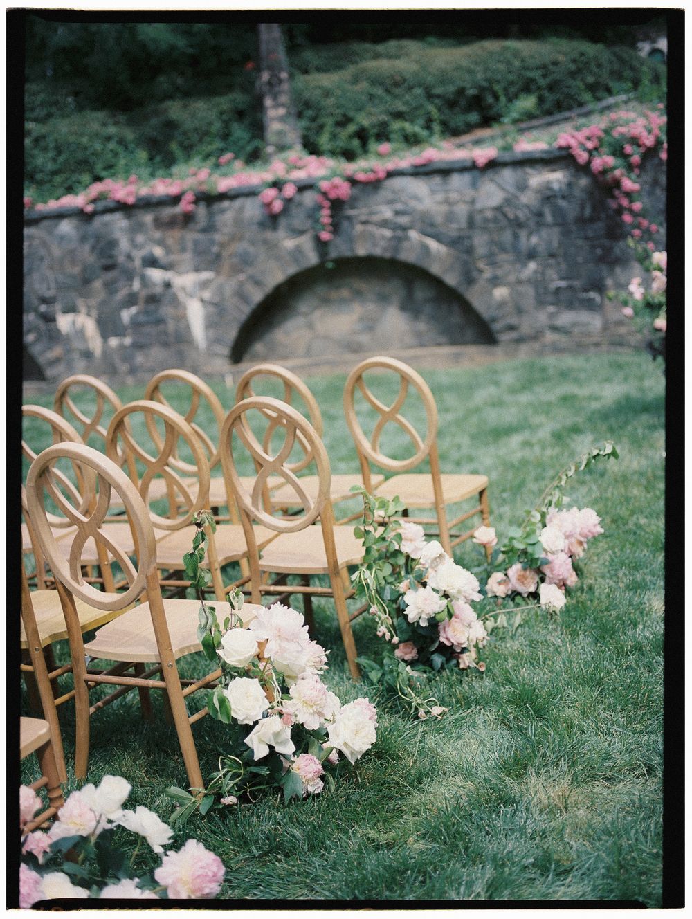 Gold chairs line a garden wedding aisle adorned with pink peonies and roses beside a stone wall.