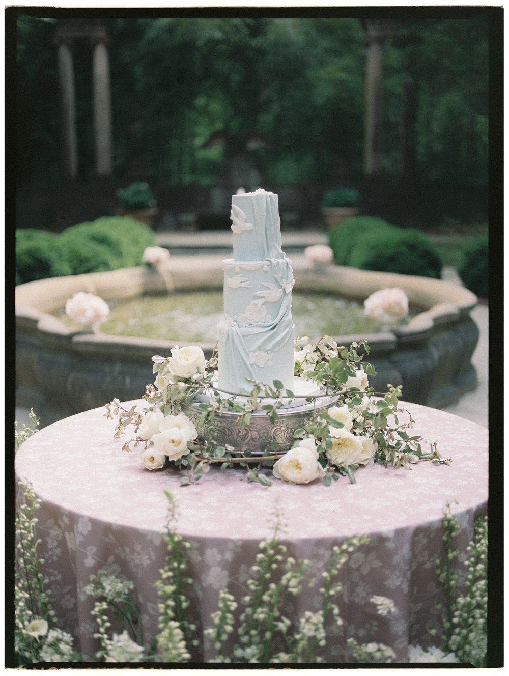 Three-tier light blue wedding cake on a round table with white floral garland, set before a garden fountain.