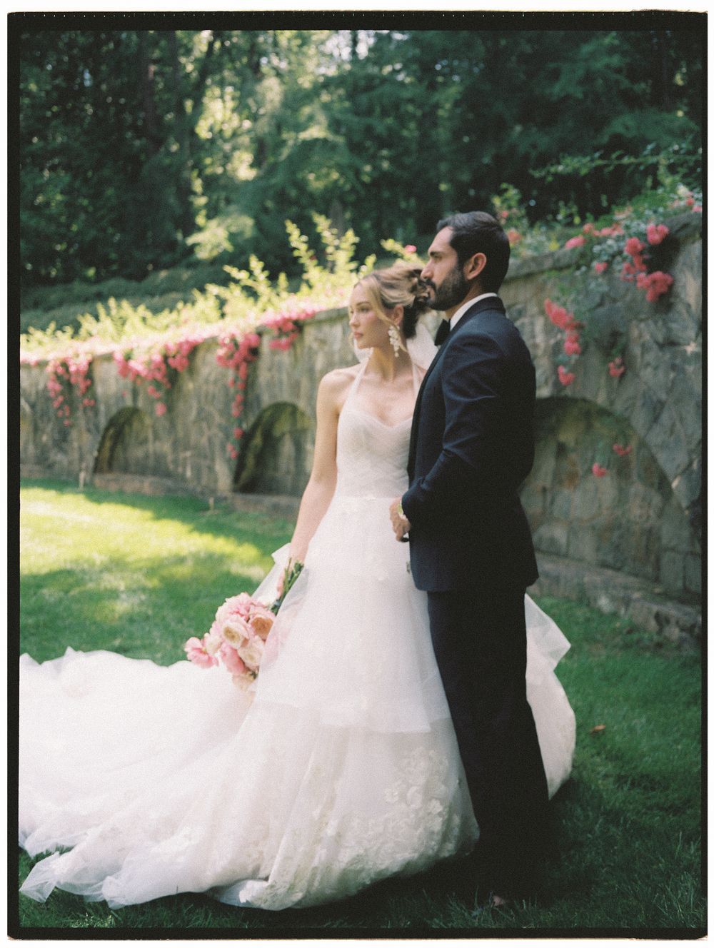 Bride in white gown holds pink bouquet beside groom in navy suit near stone wall with red roses.