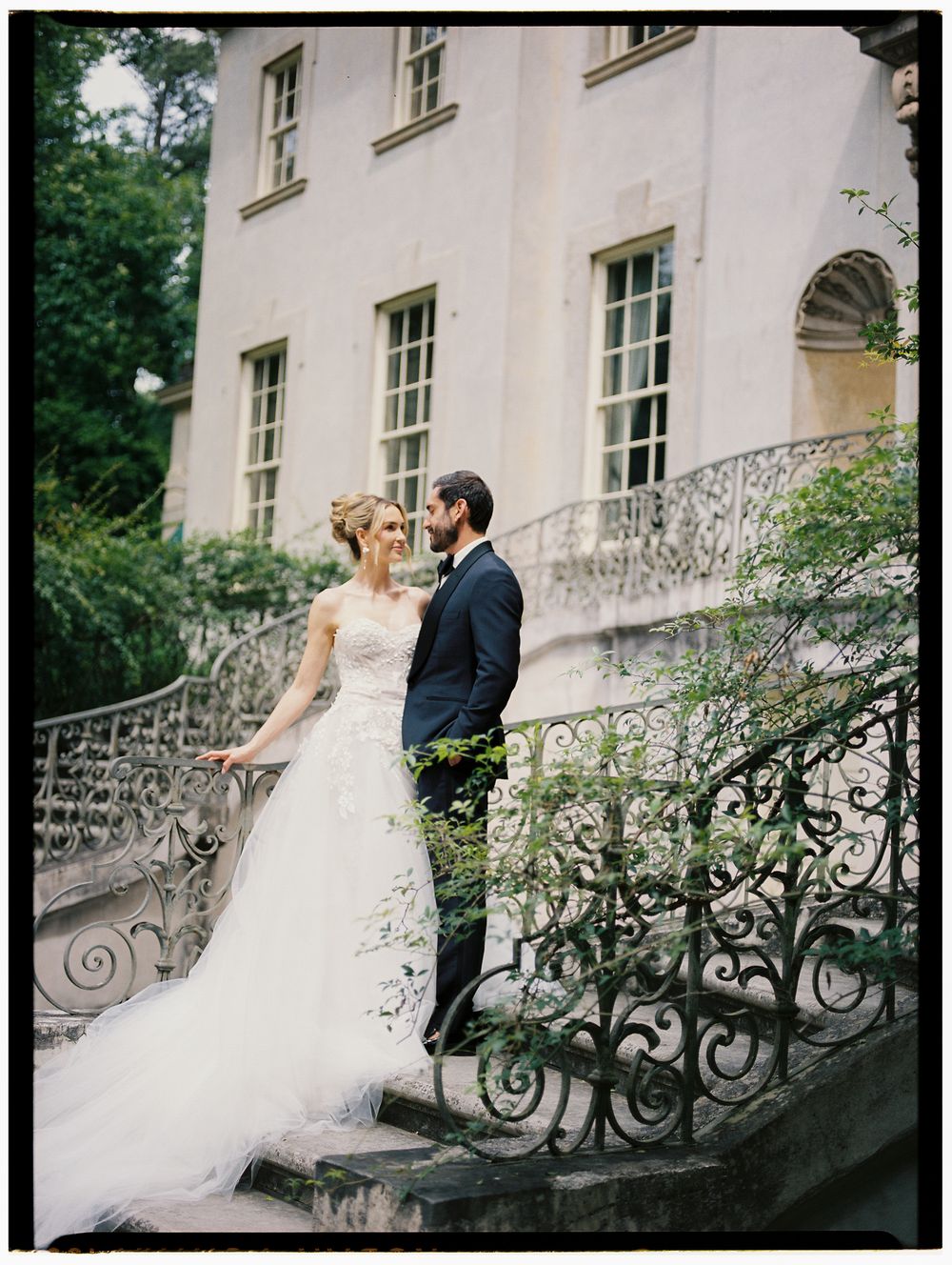 Bride and groom share a romantic moment on ornate iron staircase outside an elegant stone mansion.