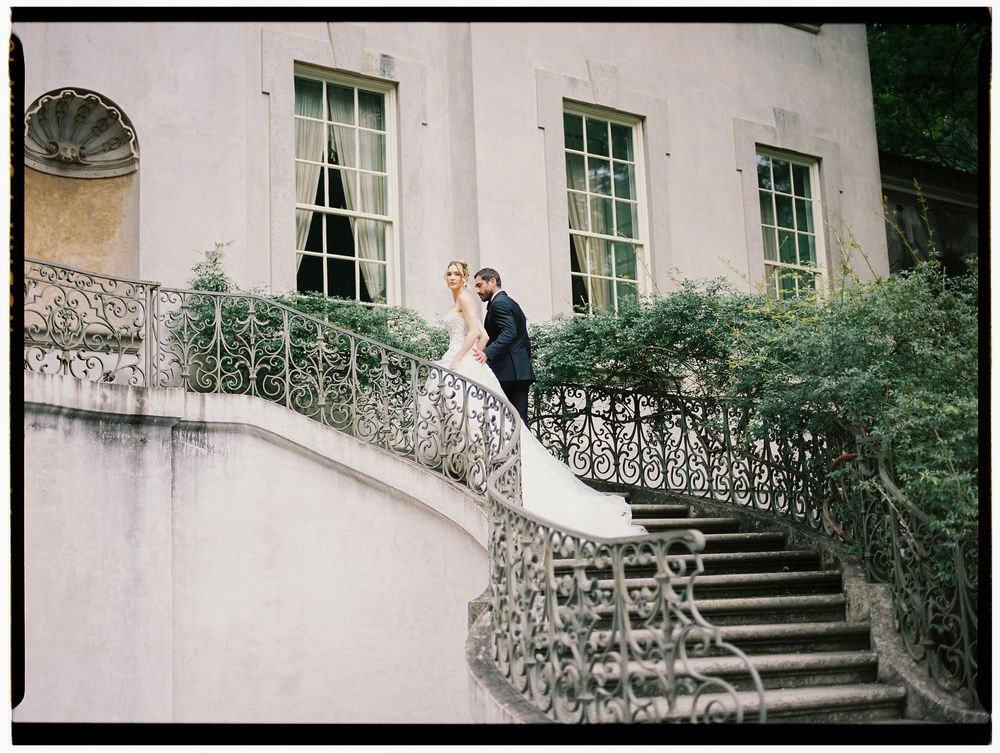 Bride and groom pose on elegant iron staircase of historic mansion surrounded by lush greenery on their wedding day.