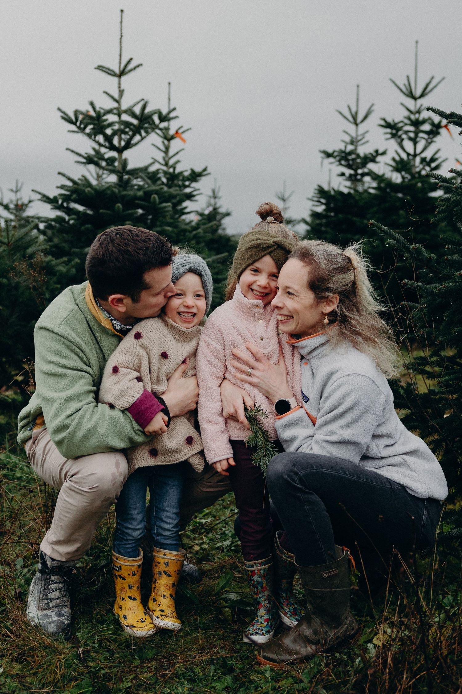 Happy family with two young girls laughing together at a Christmas tree farm on a cloudy winter day.