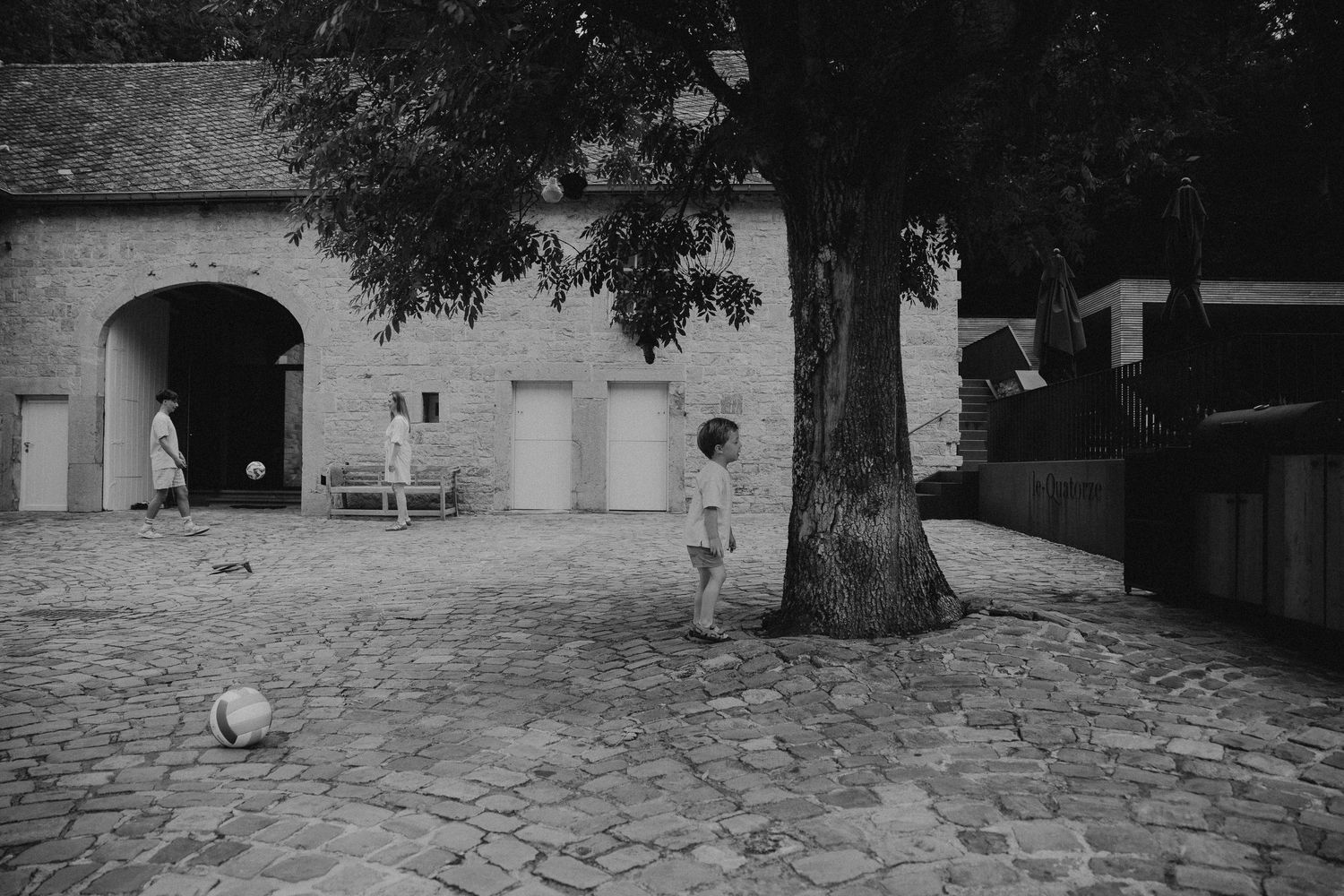 Black and white photo of a child playing near a large tree in a cobblestone courtyard beside a stone building.