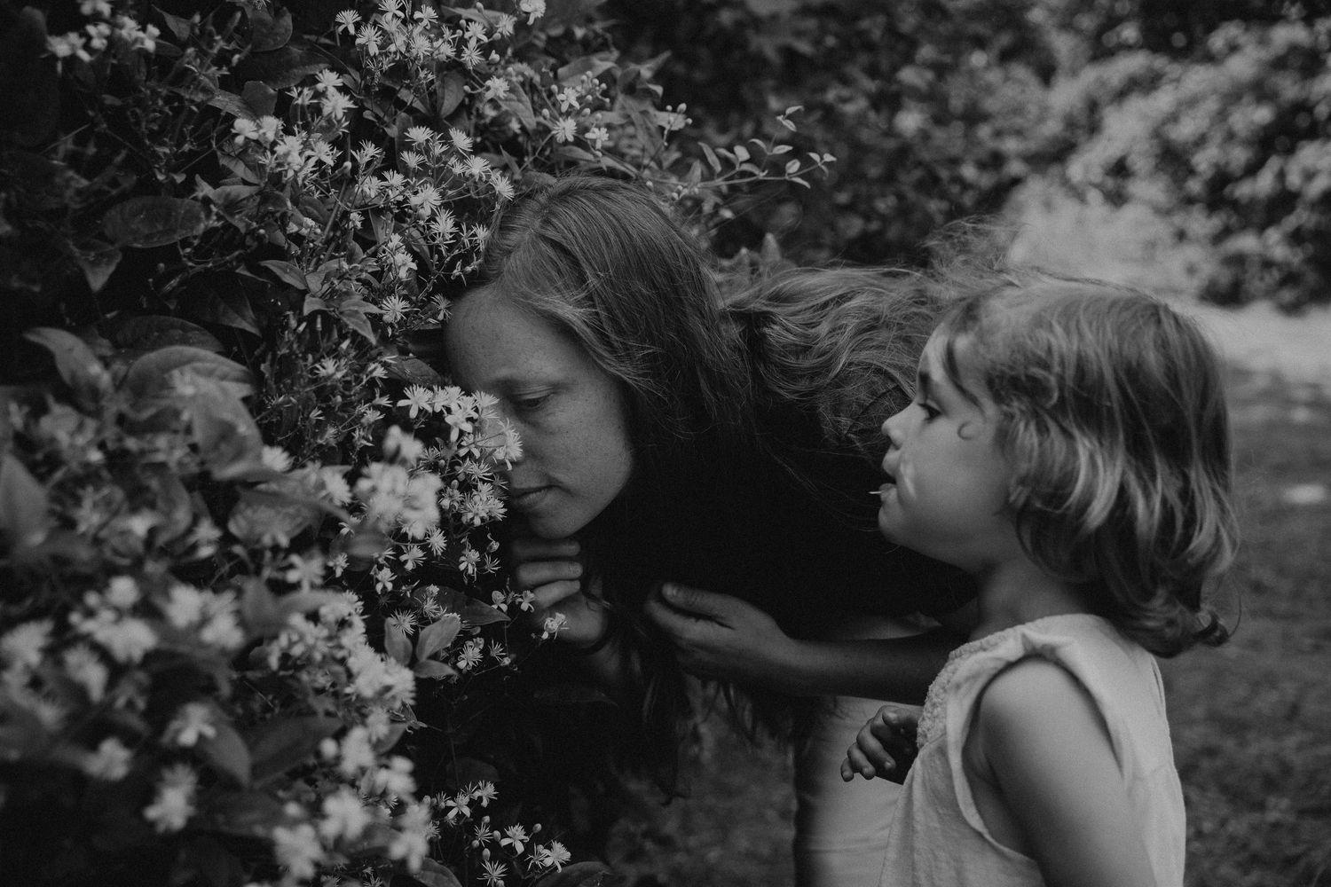 Black and white photo of a woman and young child smelling flowers on a bush in a garden.