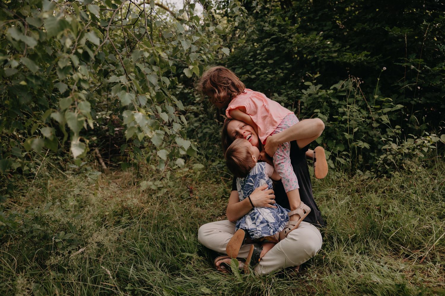 A mother sits in lush green grass hugging her two young children in a warm outdoor family embrace.
