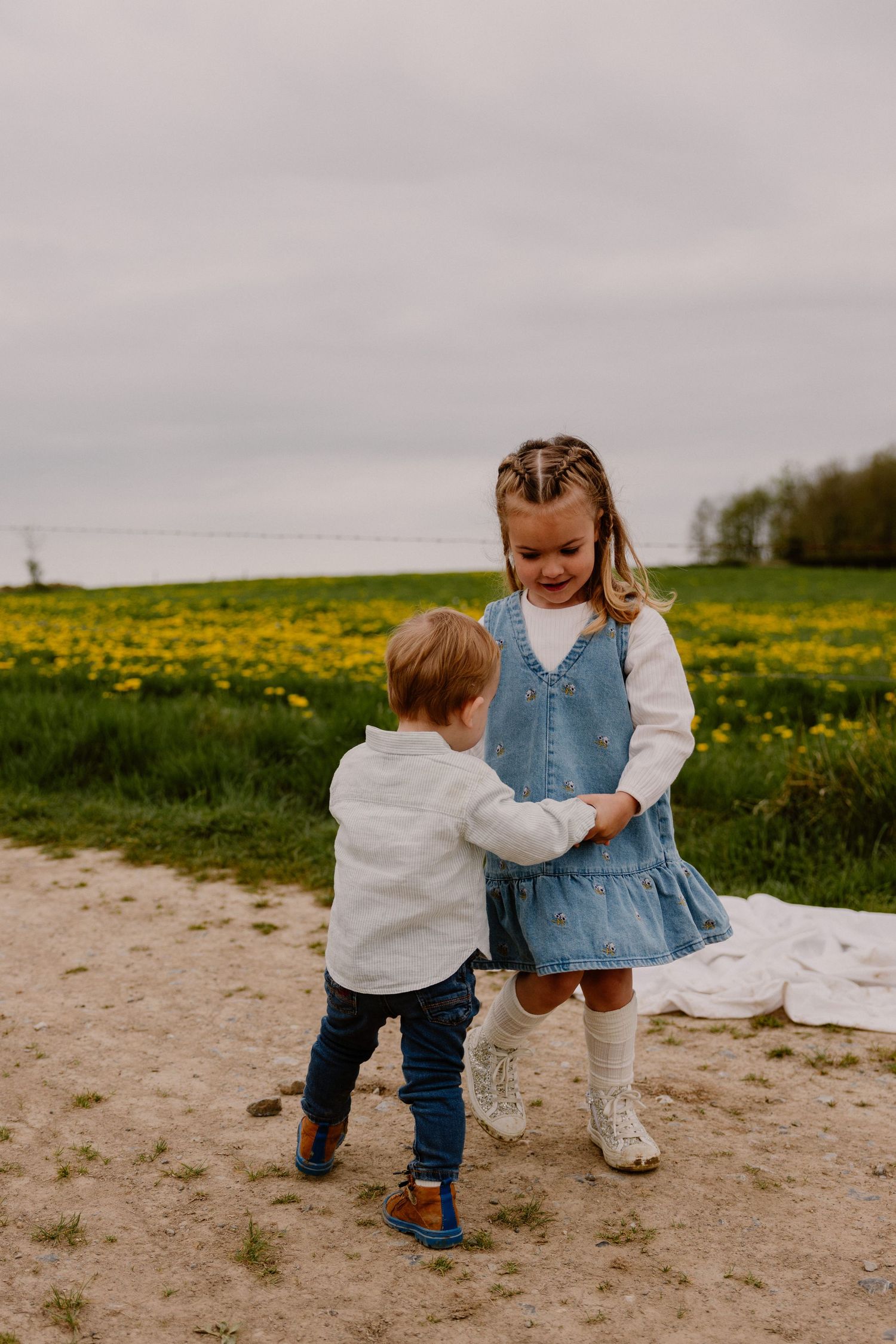 Two young children playing outdoors near a field of yellow flowers on a cloudy day.