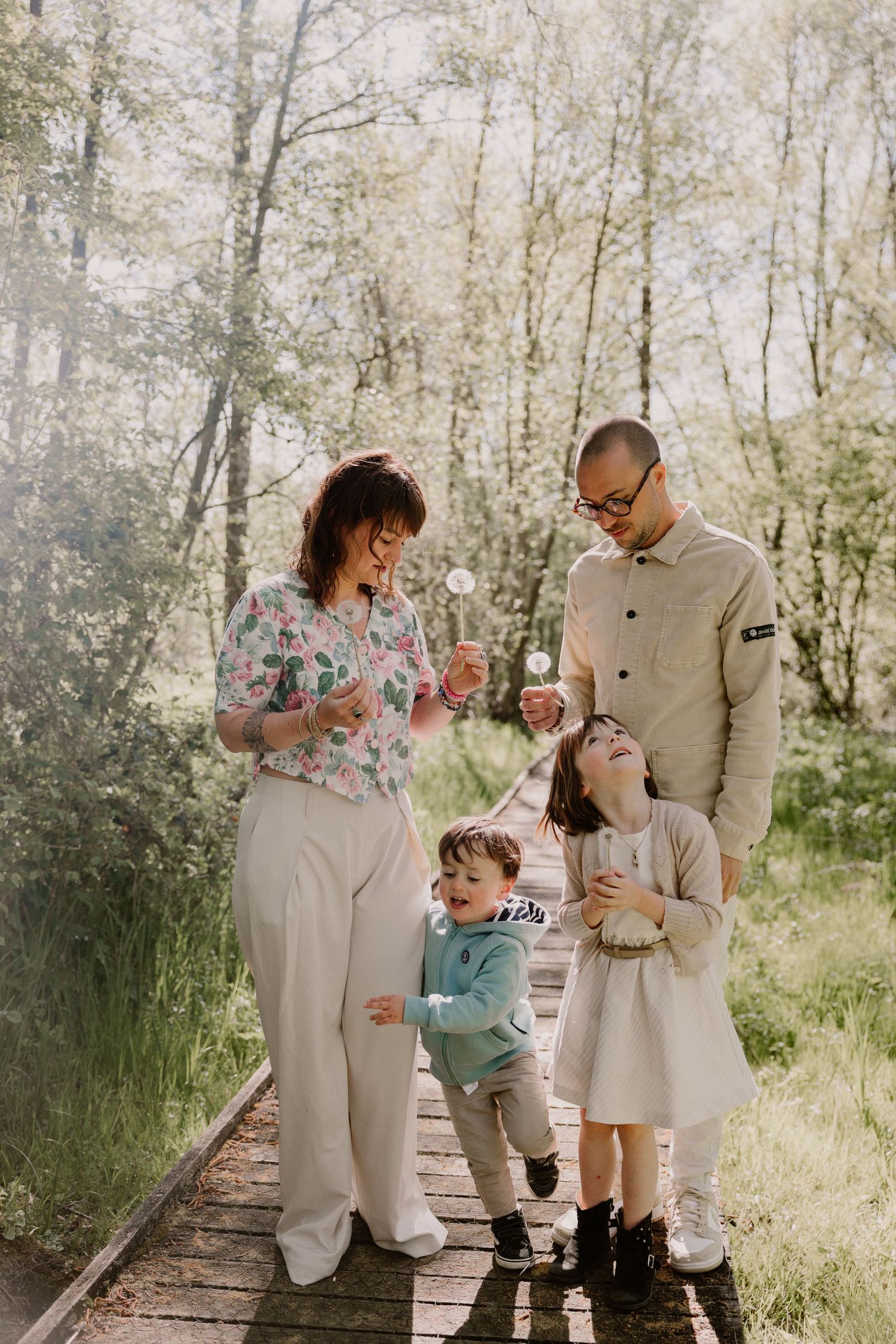 Family of four blowing dandelions on a wooden path in a sunny green forest, smiling and enjoying nature together.