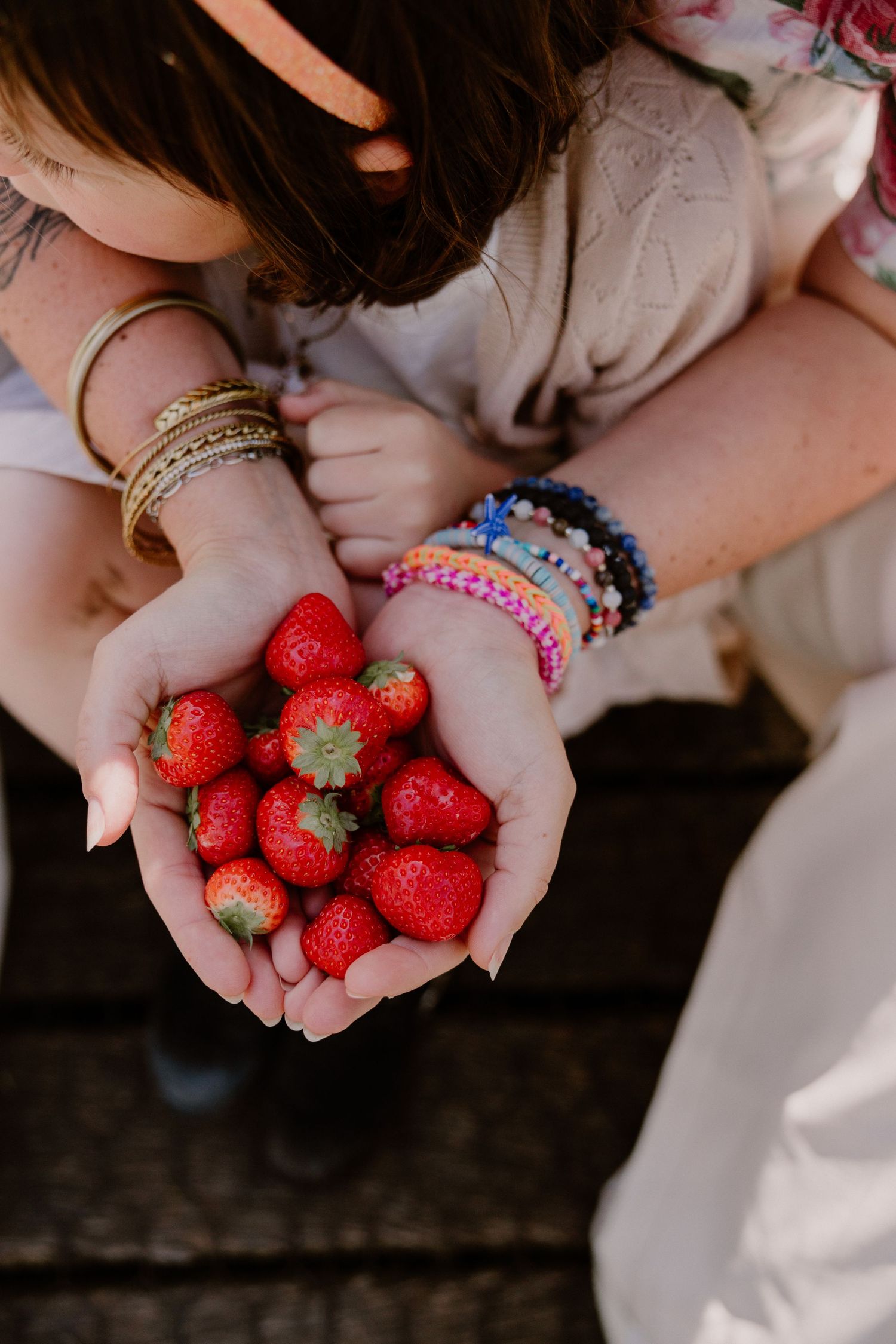 Two people holding a handful of fresh red strawberries, wearing colorful bracelets, outdoors.