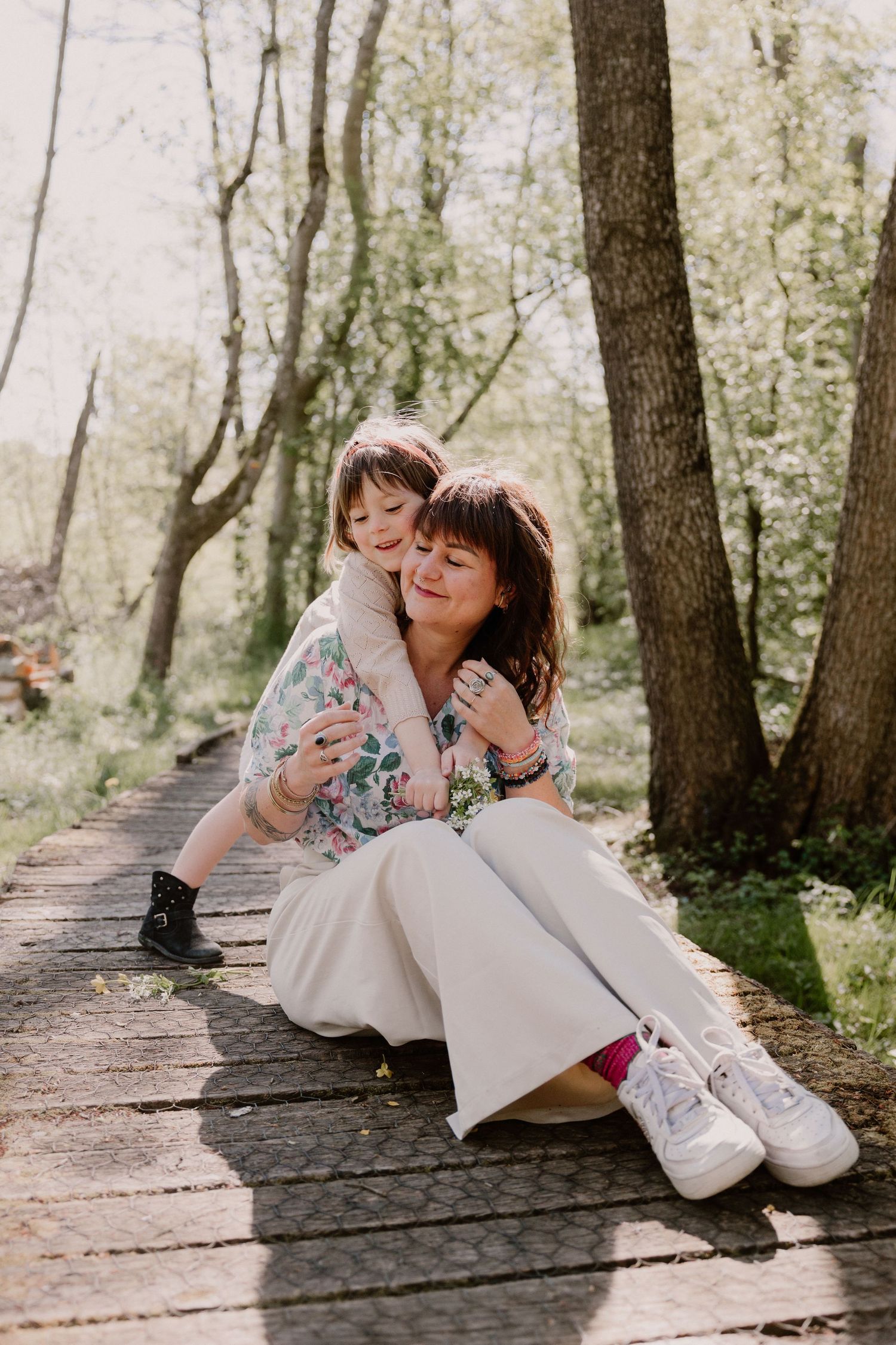A mother and young child share a tender hug on a wooden path in a sunlit forest setting.