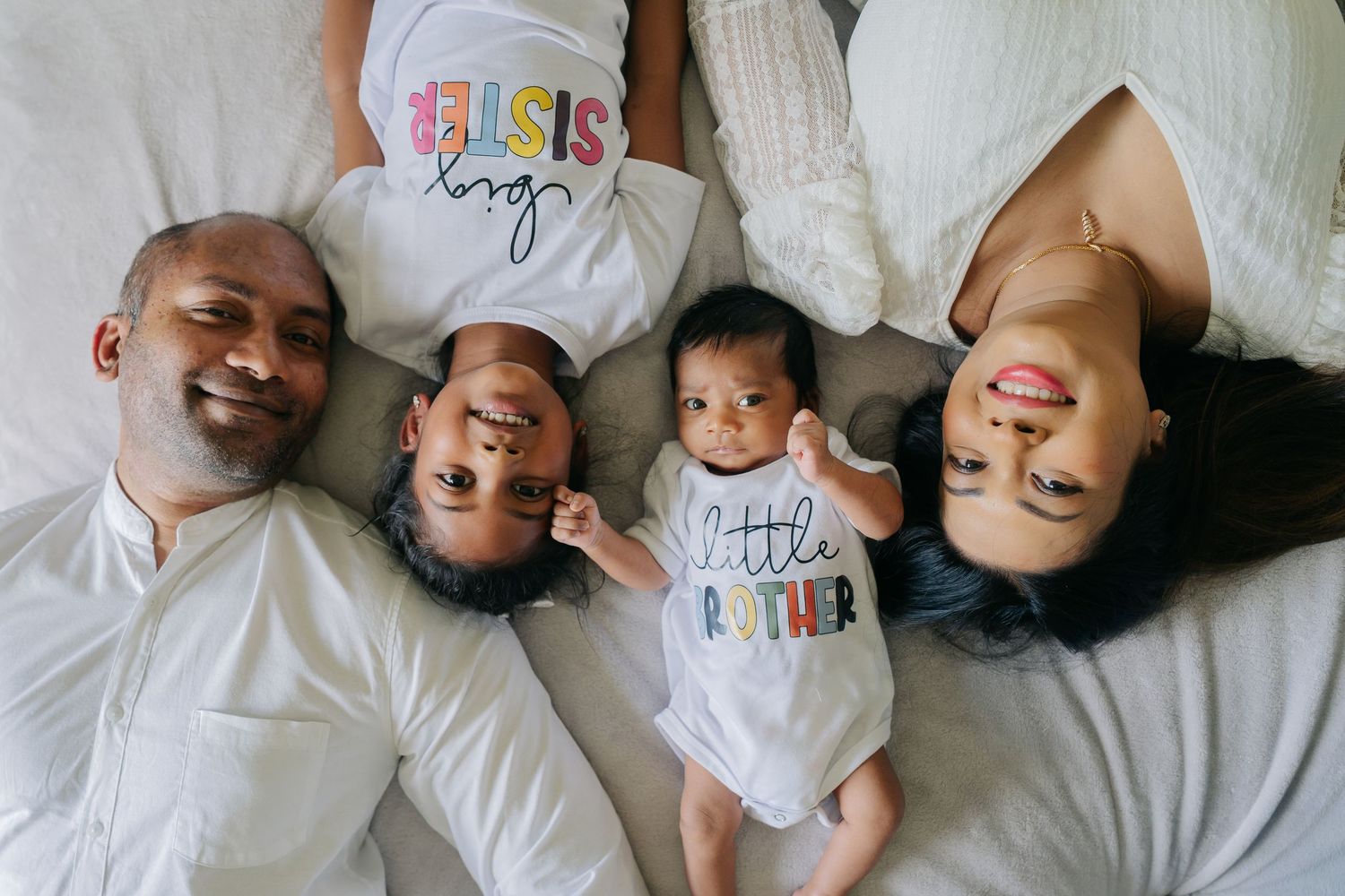 Happy family of four lying on white bed, parents smiling with two young children wearing sibling shirts.