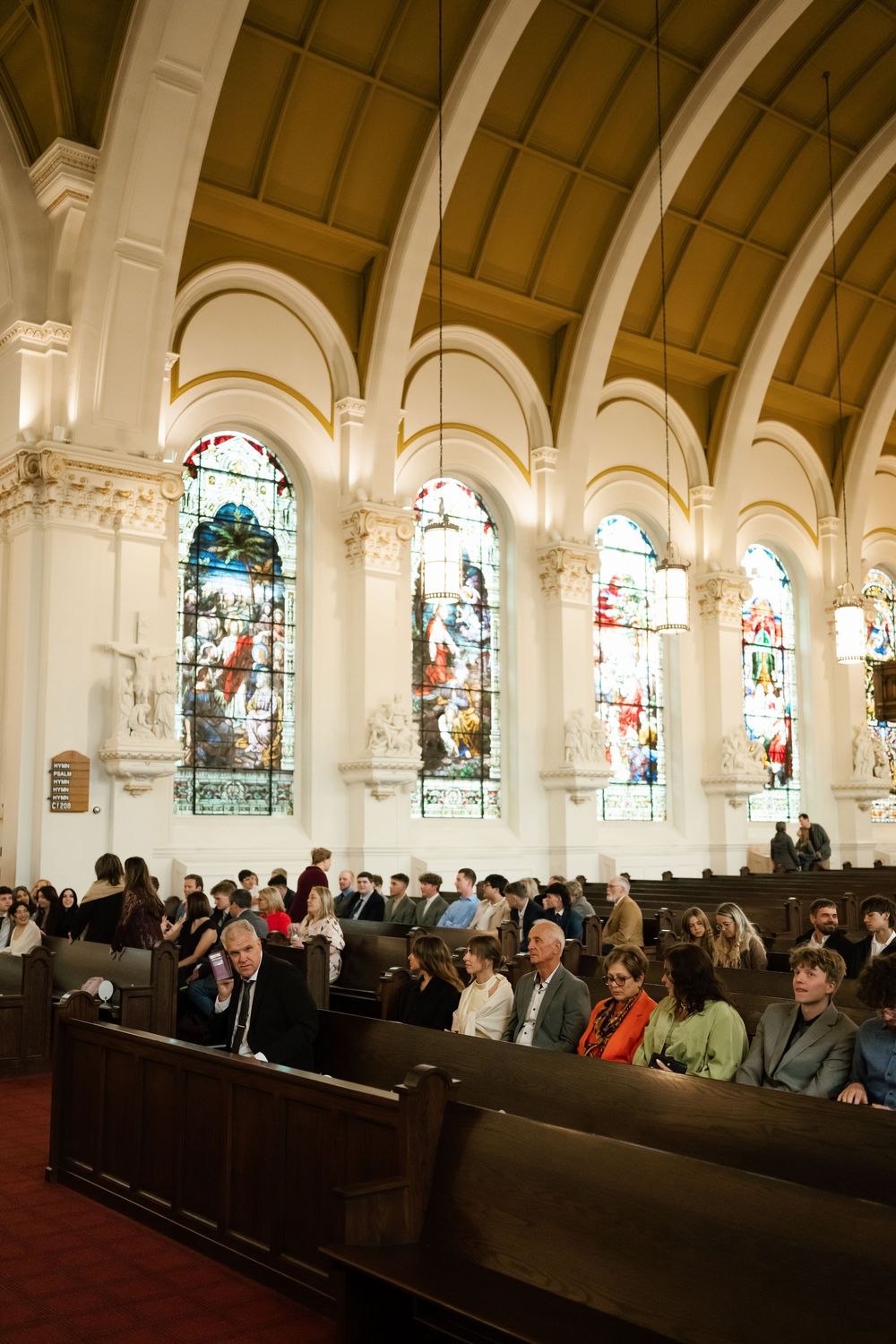 Guests seated in ornate church pews beneath arched ceilings and colorful stained glass windows during a wedding ceremony.