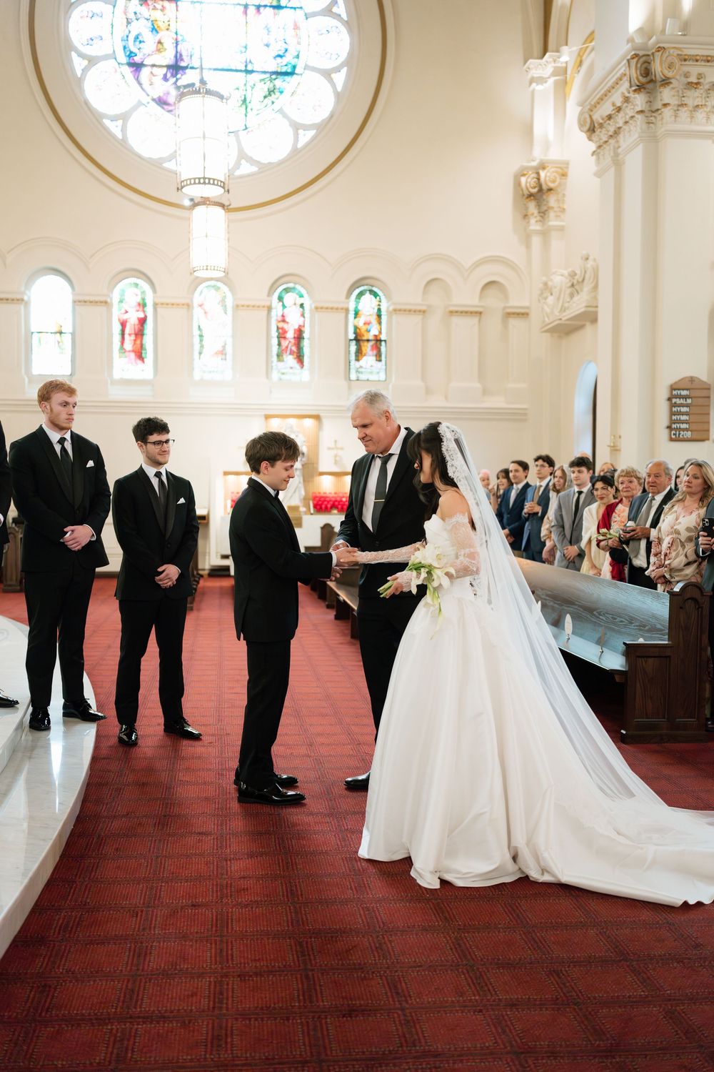Bride in white gown and veil exchanges vows with groom in black suit at elegant church wedding ceremony.