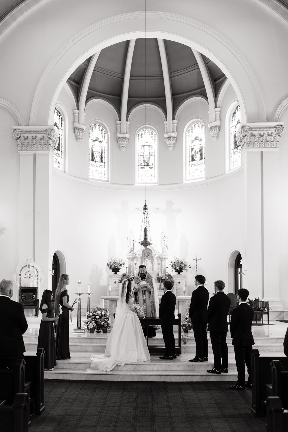Black and white photo of a bride and groom exchanging vows at a church altar with wedding party and stained glass windows.