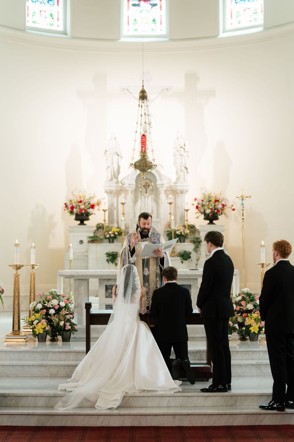 Bride and groom kneel before priest at ornate white church altar adorned with floral arrangements during wedding ceremony.
